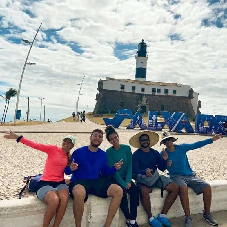 Participantes da 1ª edição sentados em frente ao letreiro de Salvador, com o Farol da Barra ao fundo