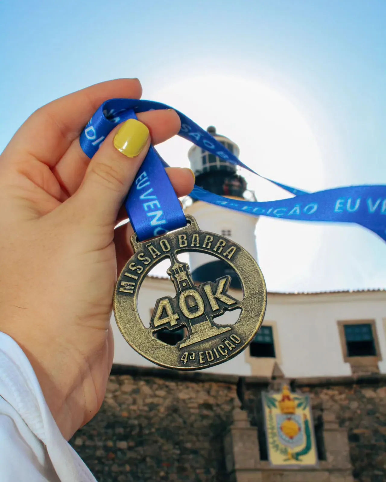 Mão segurando a medalha da 4ª edição, a primeira da Missão Barra, em frente ao forte do Farol
